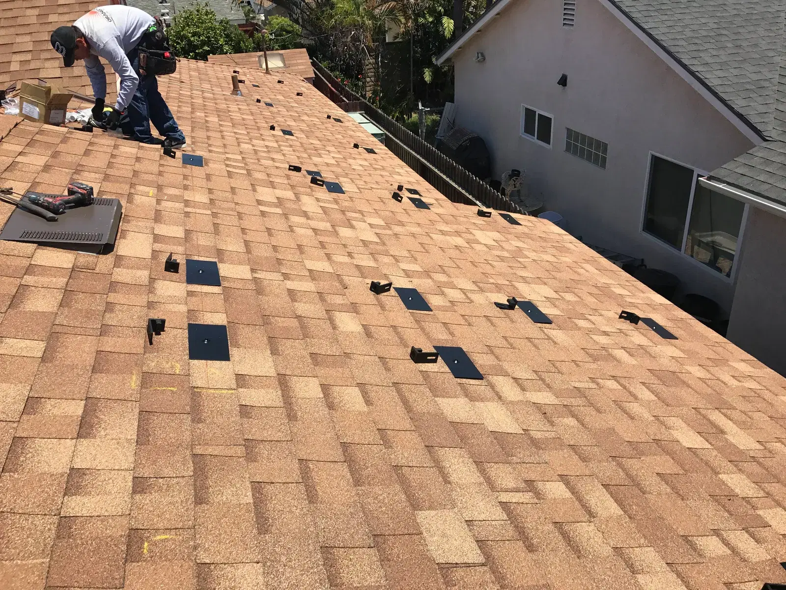 A worker wearing a hard hat and tool belt kneels on a tan asphalt-shingle roof, drilling rows of black mounting brackets in preparation for a solar panel installation.