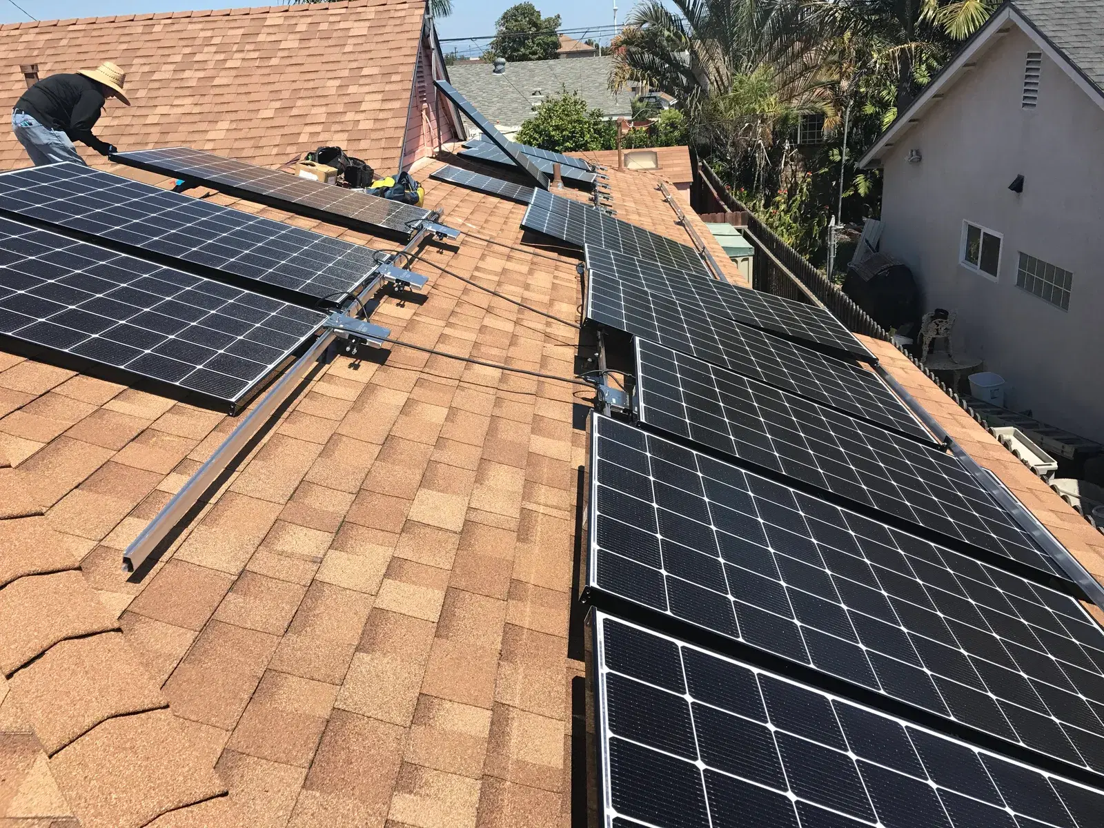 Rooftop residential solar installation with multiple rows of black rectangular solar panels on tan asphalt shingles, a worker at the upper-left adjusting a panel.
