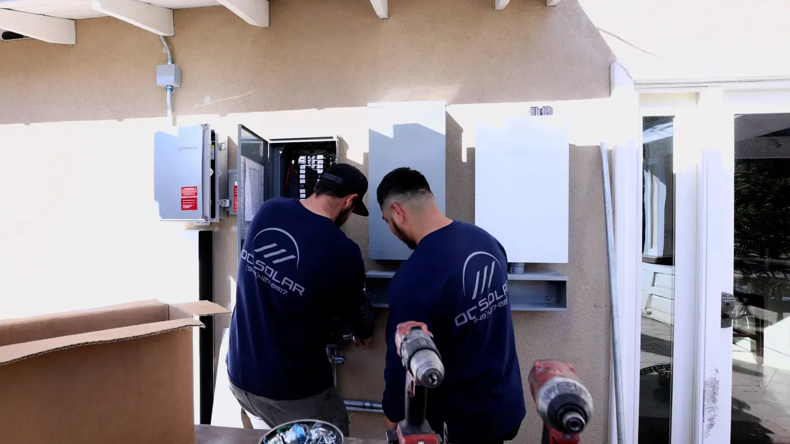 Two OC Solar technicians in navy shirts drilling and wiring grey electrical panels mounted on a stucco wall.