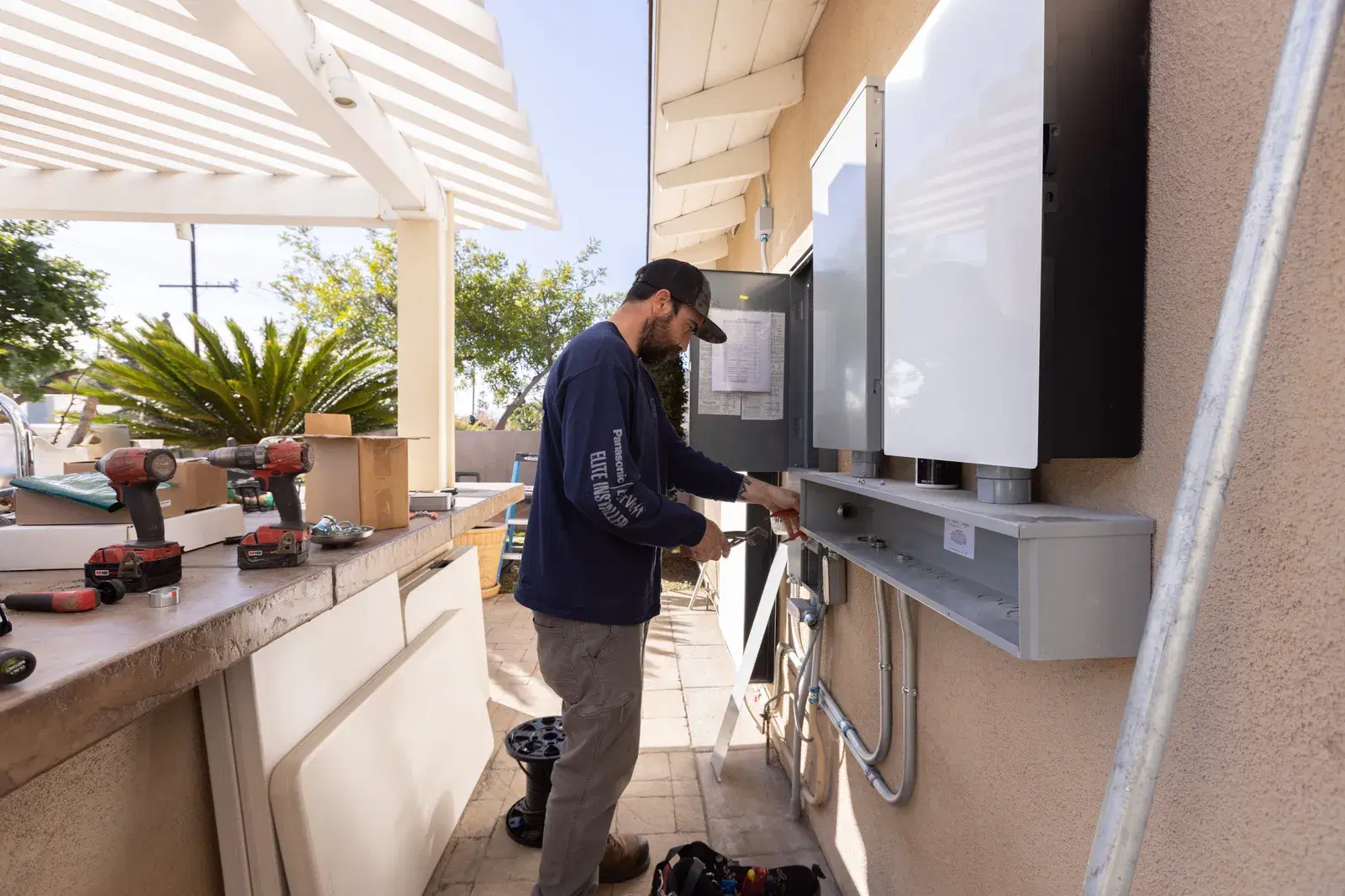 A technician in a navy long-sleeve shirt wiring electrical equipment and conduit on a stucco exterior wall, drills on a ledge.