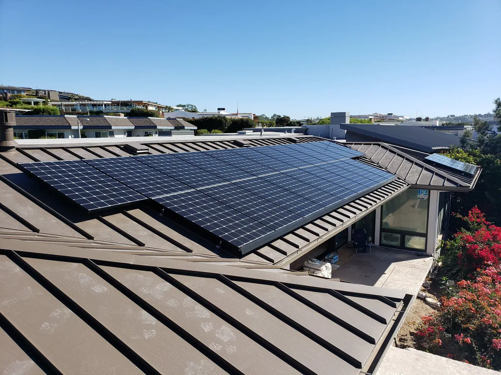 Solar panels mounted on a bronze standing-seam metal roof of a hillside home under clear blue sky.