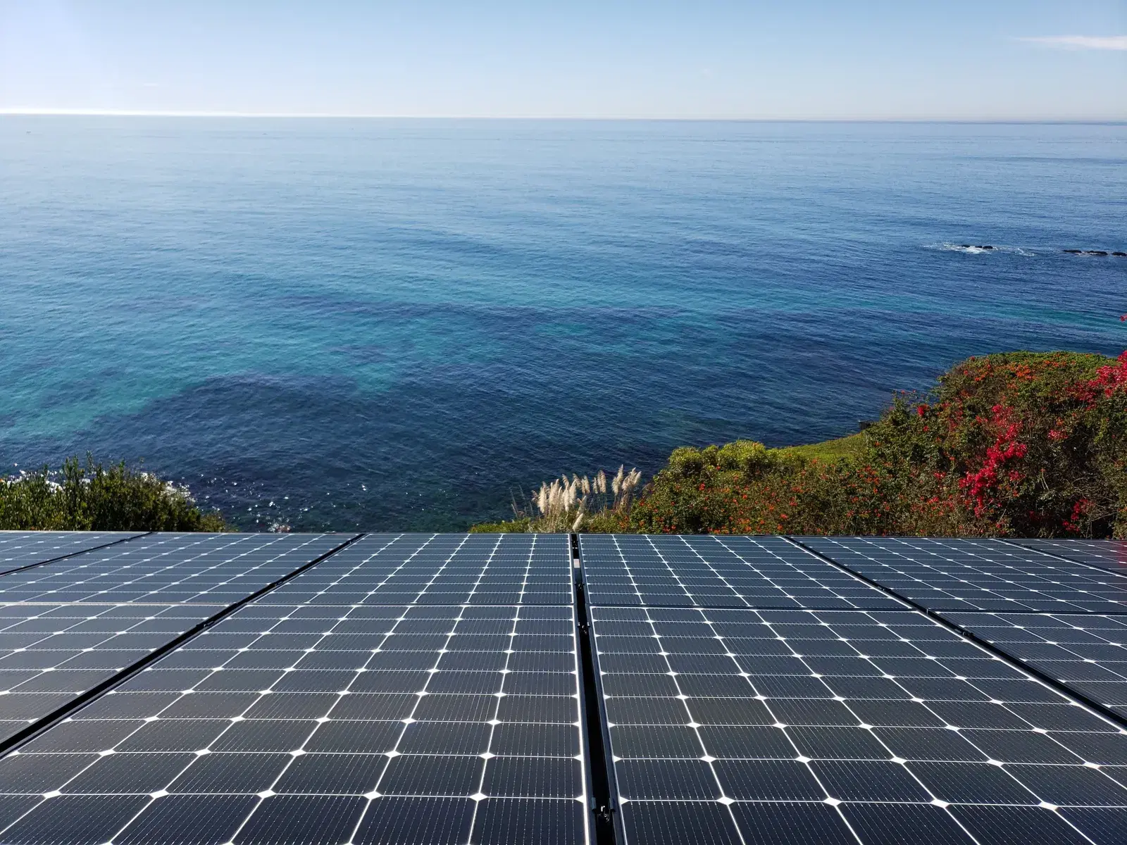 Close-up of solar panels with a vivid blue-green ocean and coastal vegetation in the background.