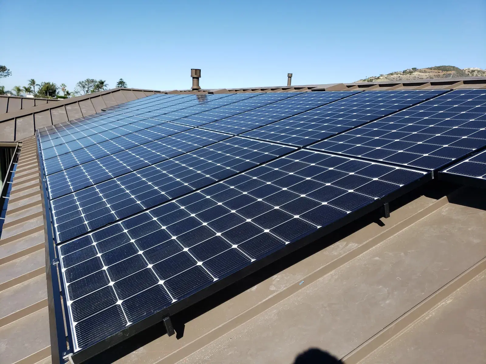 Solar panels with white-gridded cells mounted on a brown metal roof beneath a clear blue sky.
