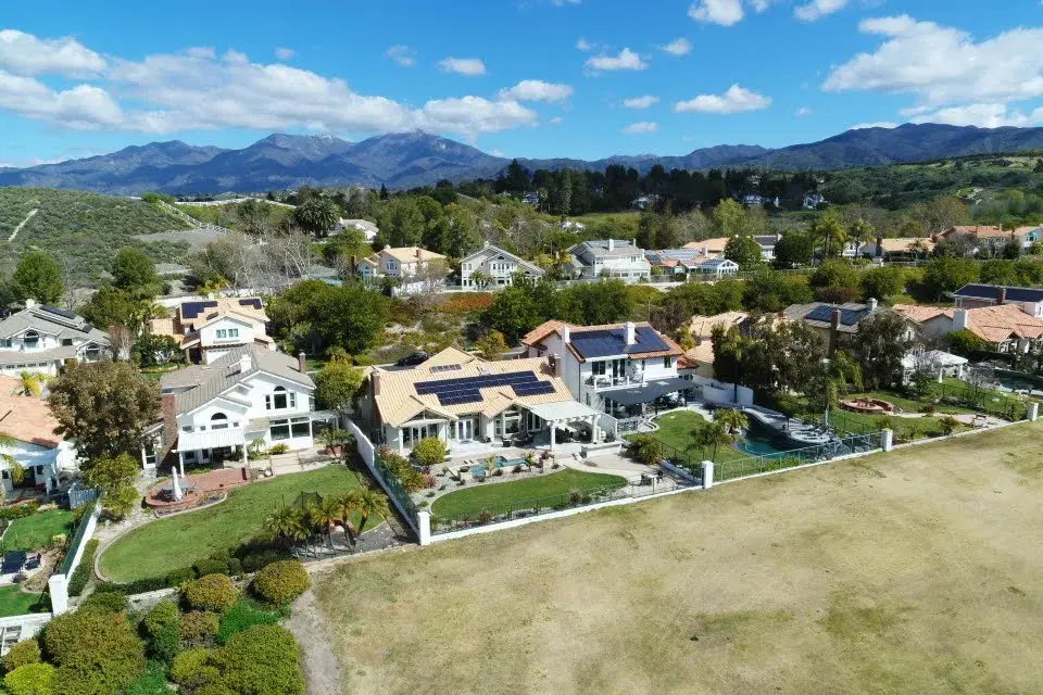 Aerial view of a hilltop neighborhood where a home displays a rooftop solar array, with mountains in the distance.