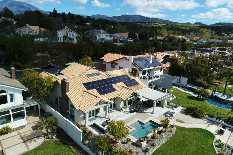 Aerial view of a large Spanish-tile-roof home with rooftop solar panels, a swimming pool and patio below, and mountains beyond.