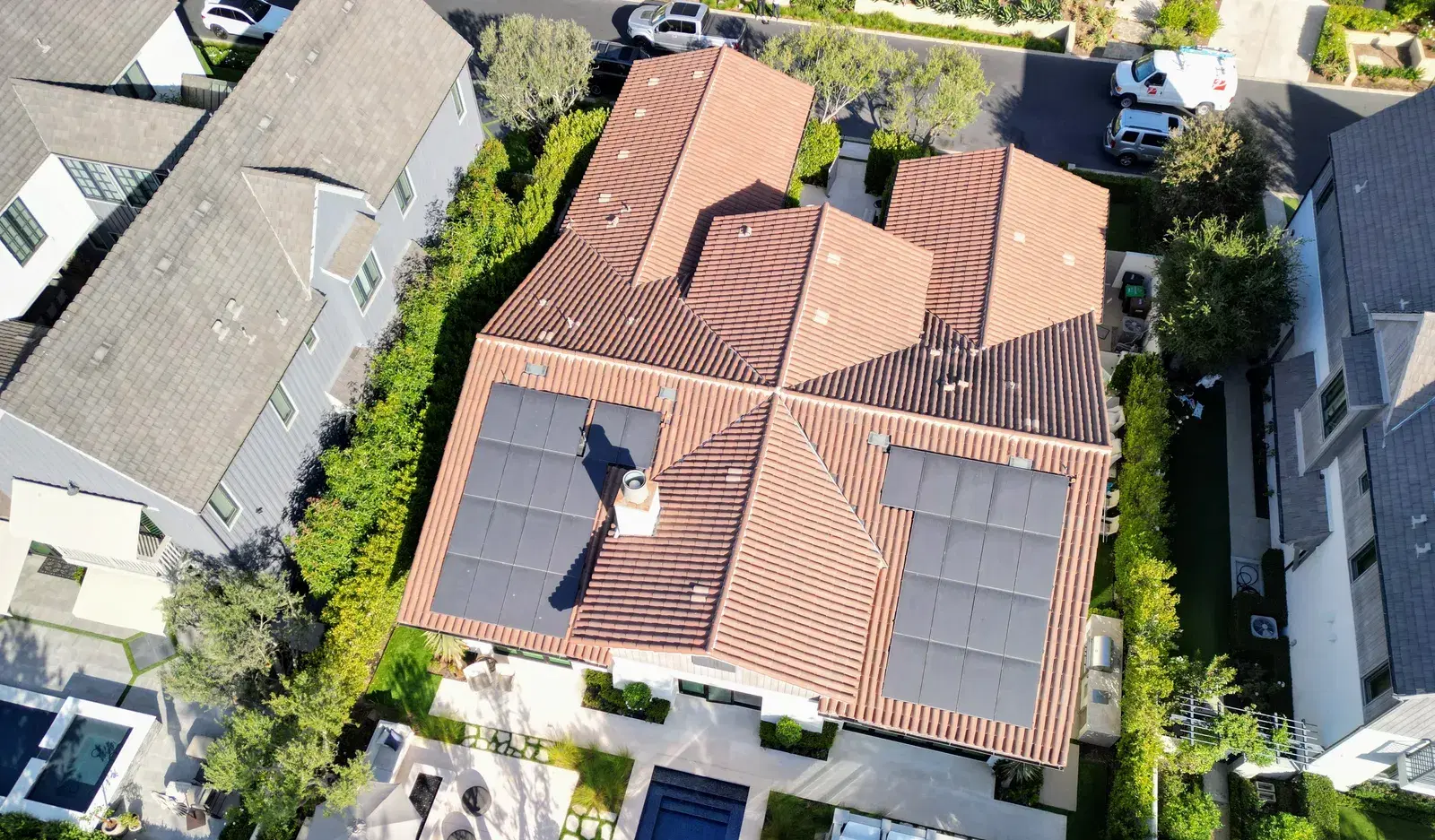 Drone shot looking down on a red clay-tile residential roof with two rectangular arrays of black photovoltaic panels, surrounded by green hedges and trees.
