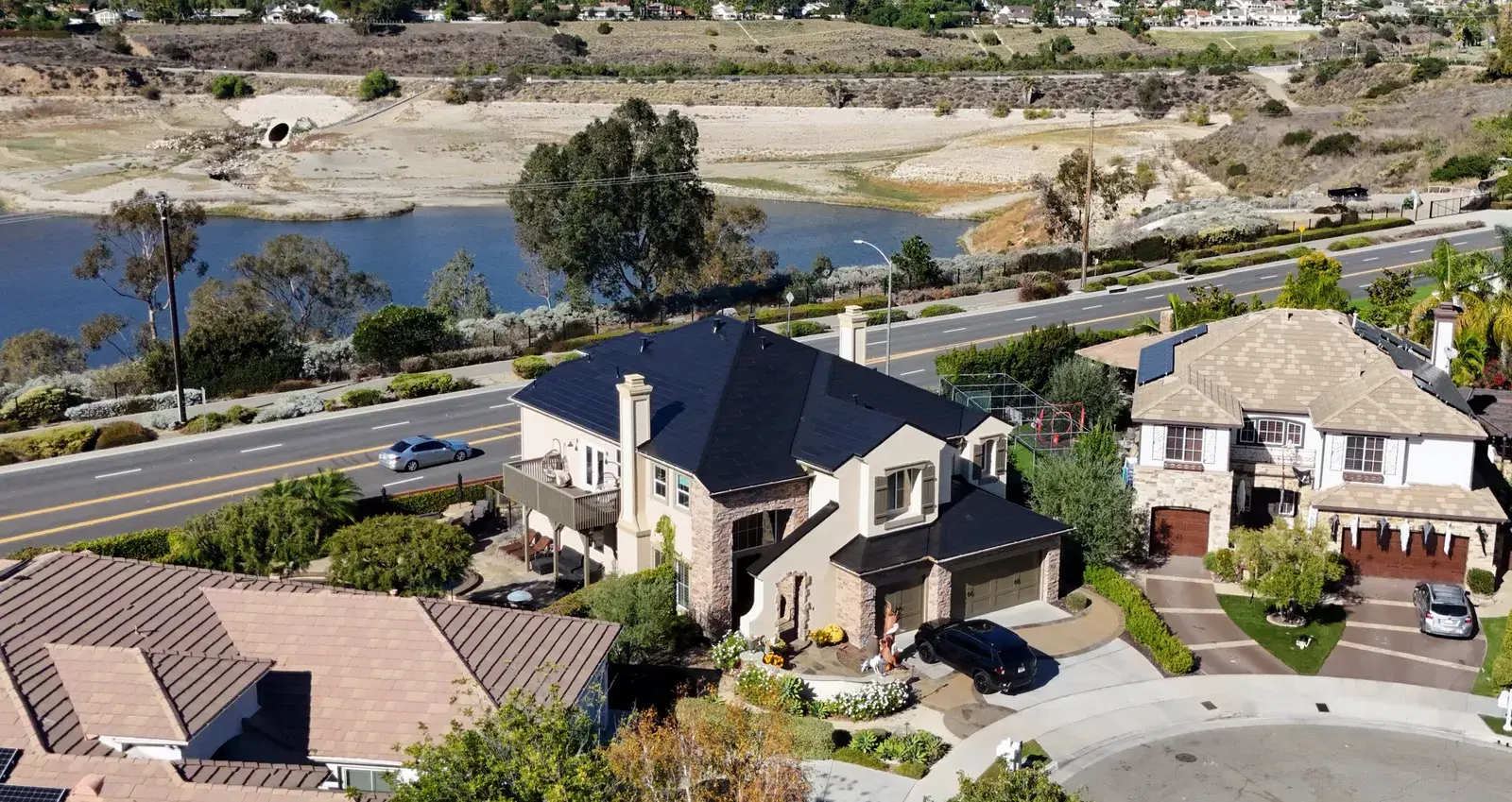 Drone shot of a two-story Southern California home with rectangular solar panels mounted on its dark roof, set in a neighborhood beside a reservoir.