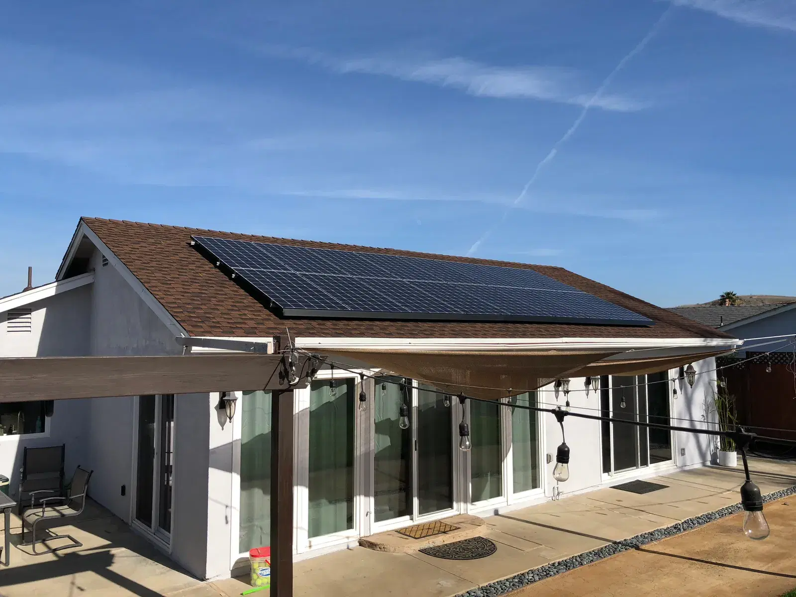 Rear of a single-story home with a rooftop solar array, patio, and string lights under a blue sky.