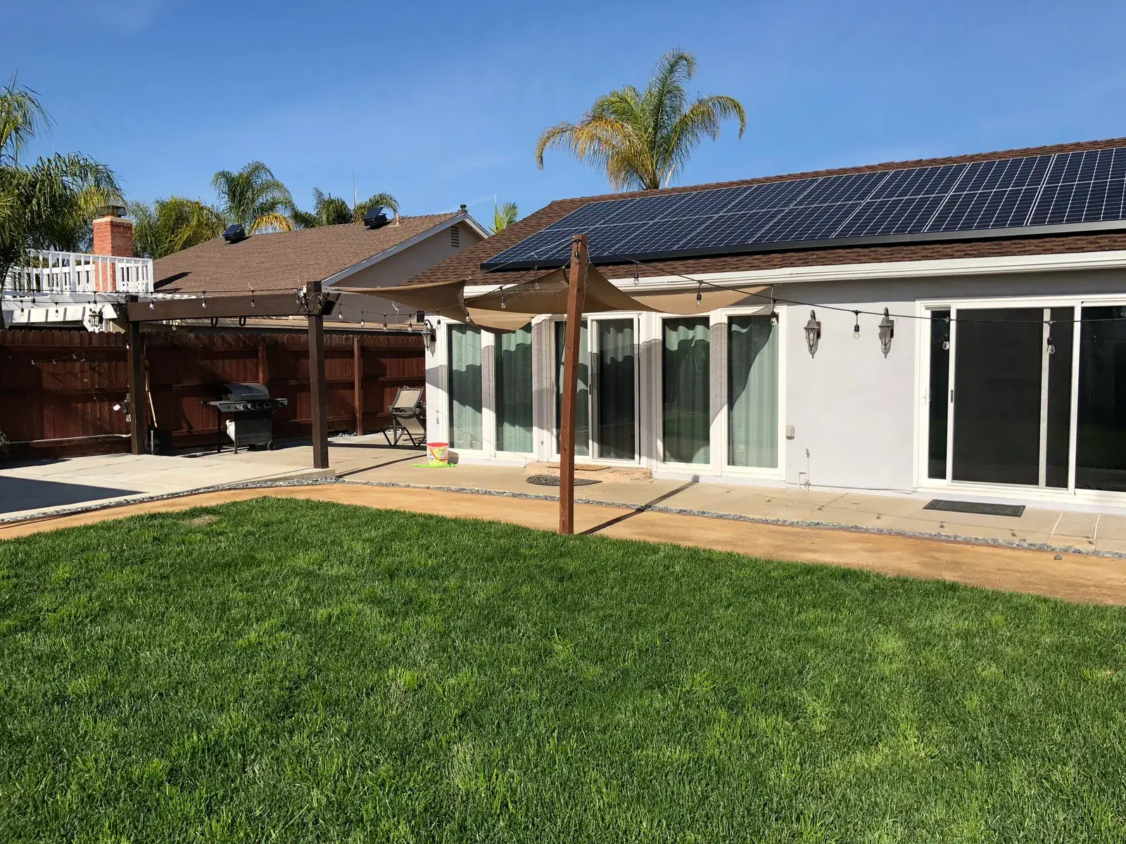Backyard with a lawn and patio looking toward a house with a rooftop solar panel array.