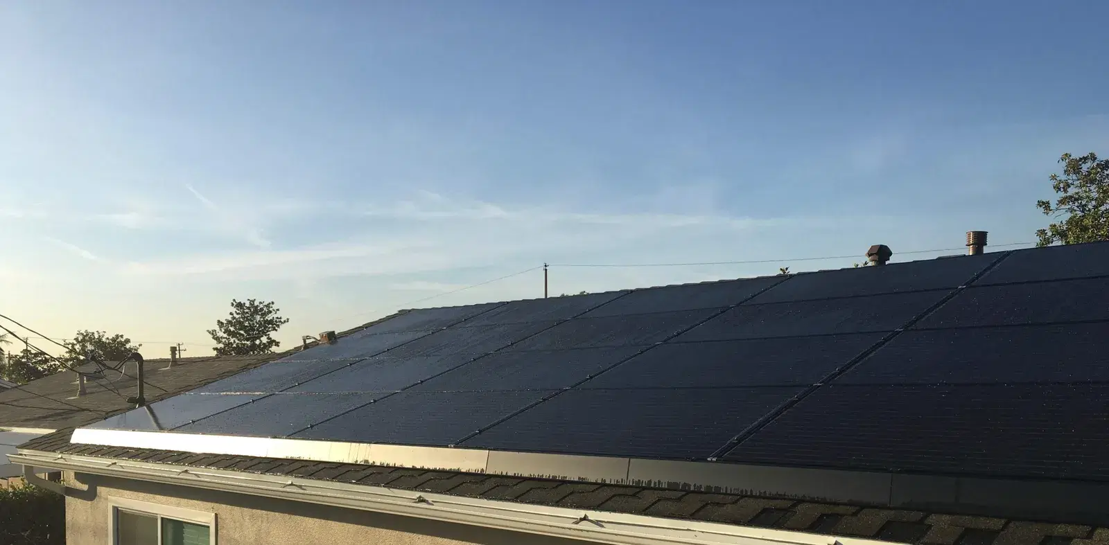 A finished black solar panel array on a residential roof seen from the side with roof vents and trees nearby.