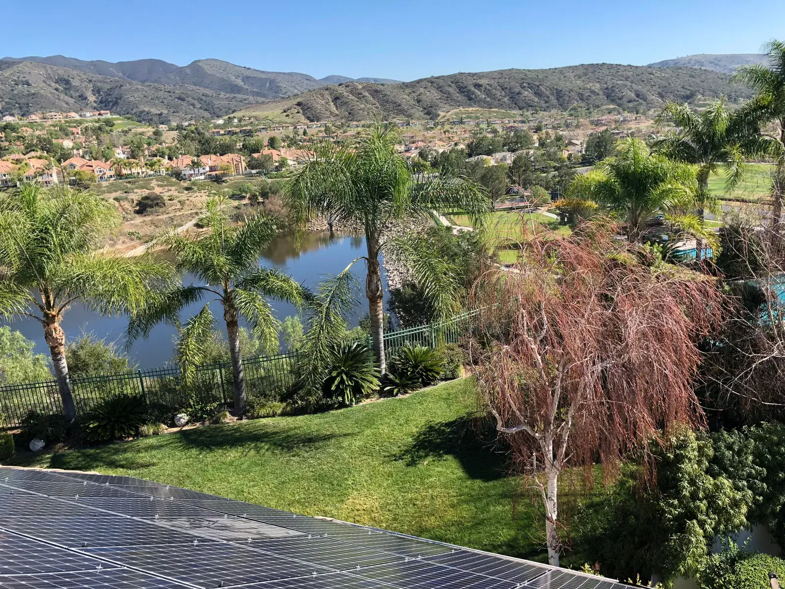 Edge of a rooftop solar panel array in the foreground with a scenic lake, palm trees and mountains beyond.