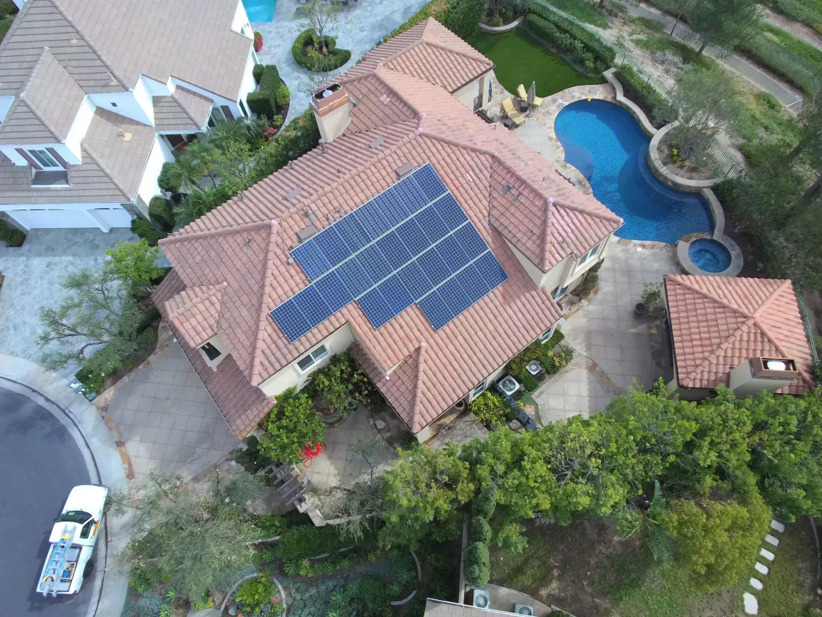 Drone photo looking down on a large tile-roofed home with a blue solar panel array, a kidney-shaped pool, and lush landscaping.
