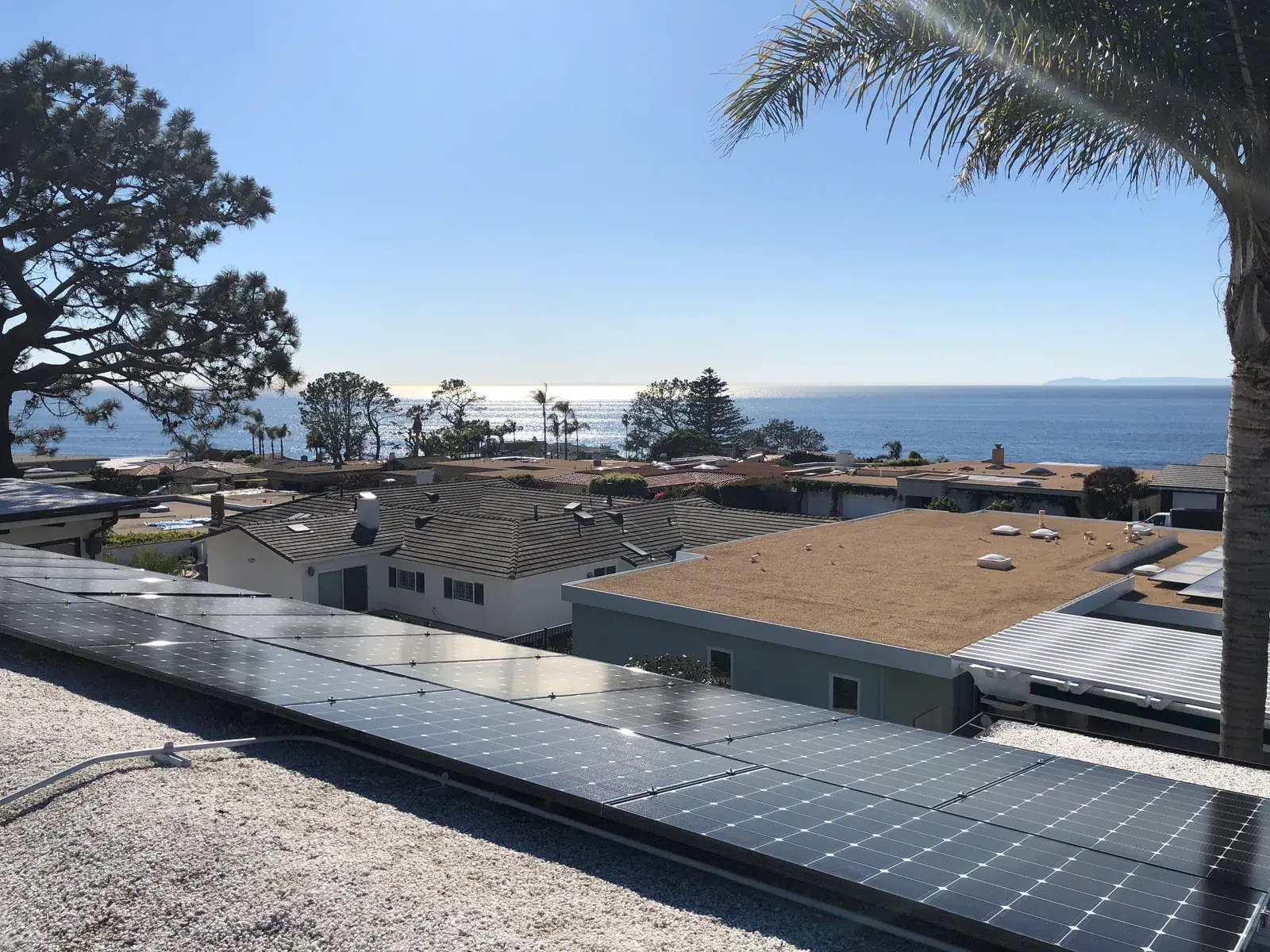 Dark solar panels mounted on a flat roof in the foreground with coastal homes and the sparkling Pacific behind.