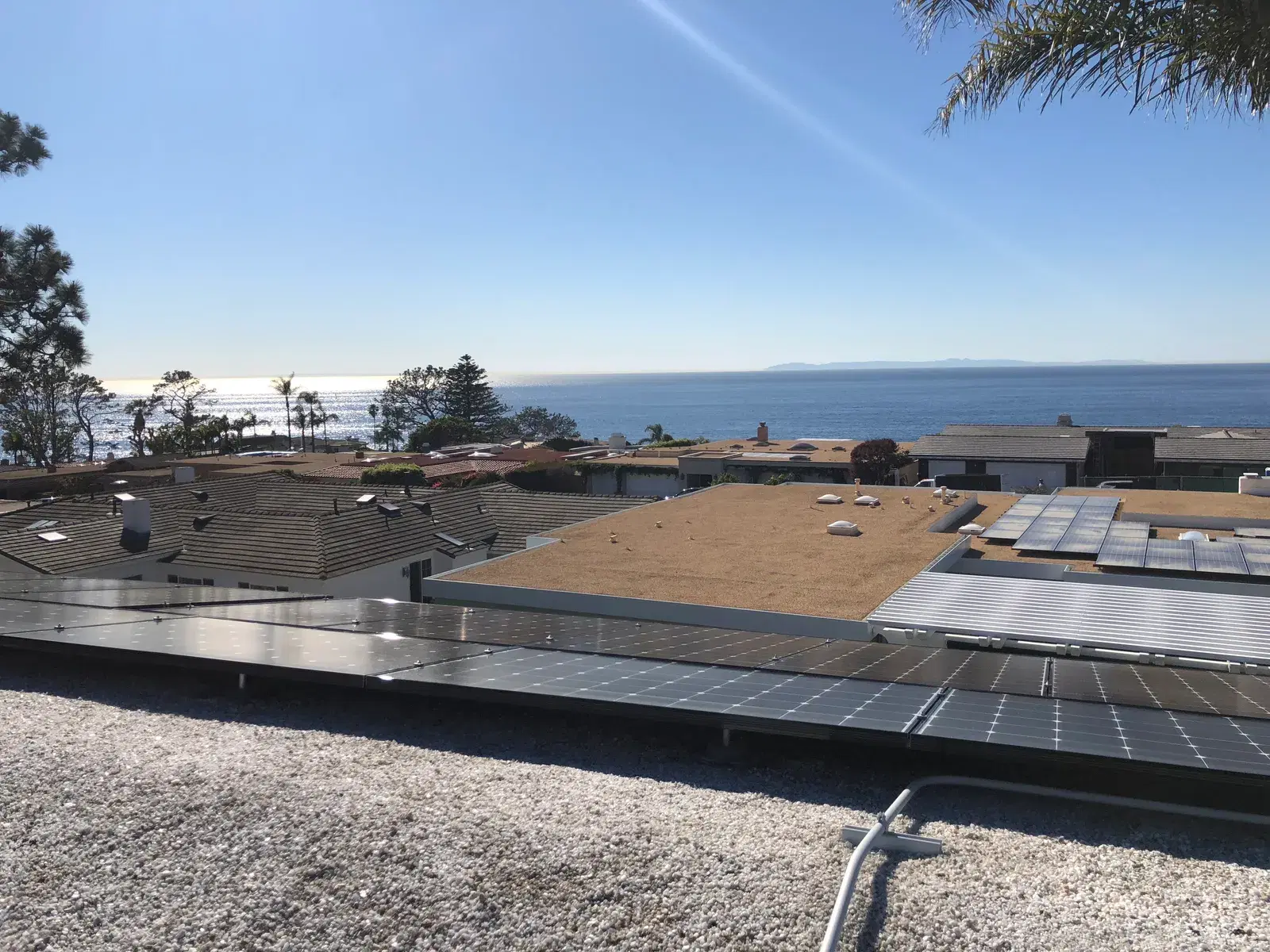 Solar panels on a gravel-ballasted flat roof in the foreground with coastal rooftops, the sea, and an island beyond.