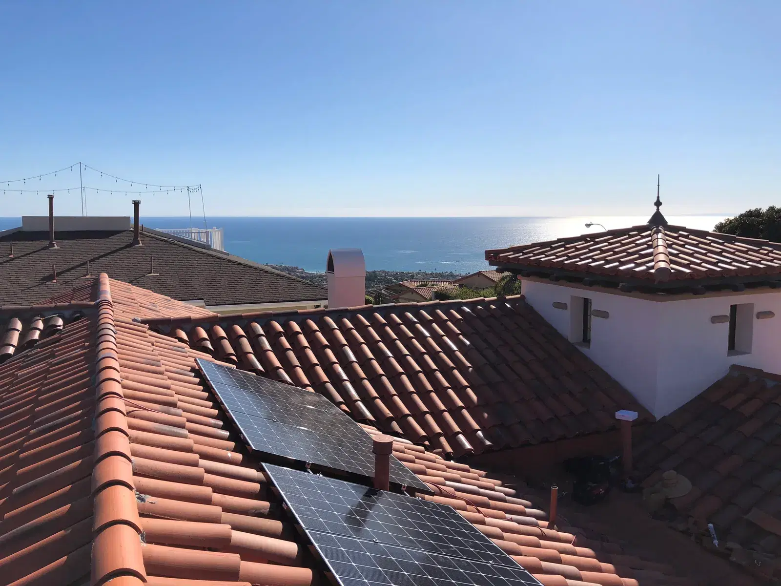 Rectangular solar panels mounted on a red clay tile roof above a coastal view.