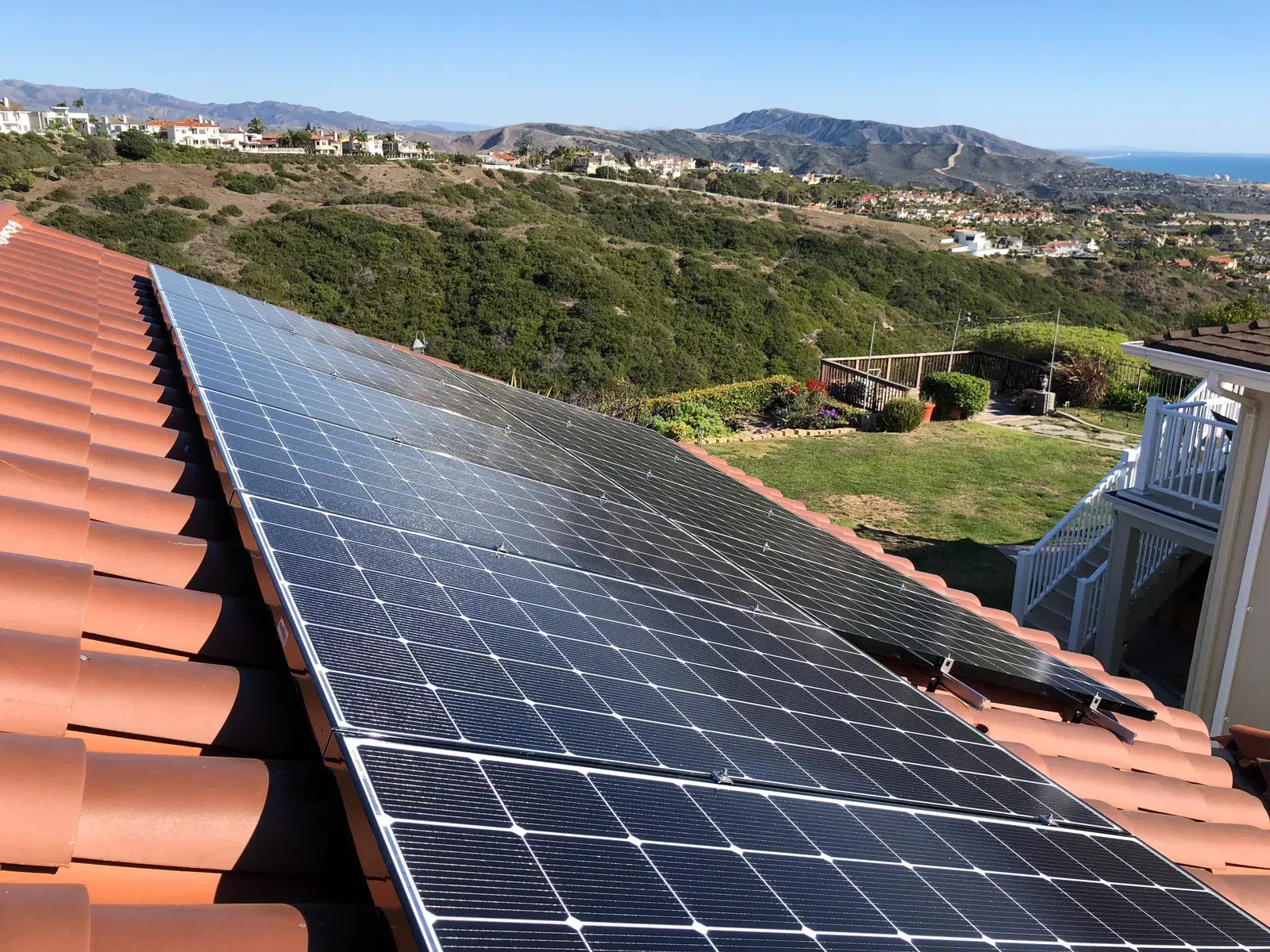 Rows of solar panels on a red tile roof overlooking green hills and the ocean.
