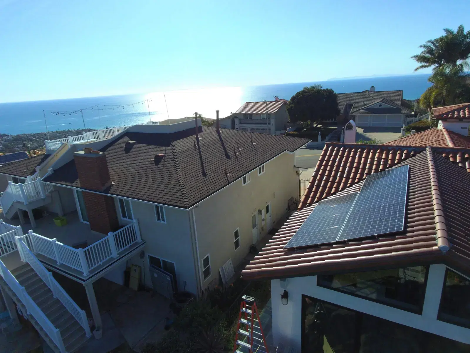 Drone shot of a hillside home with rectangular solar panels mounted on a red clay-tile roof overlooking the ocean.