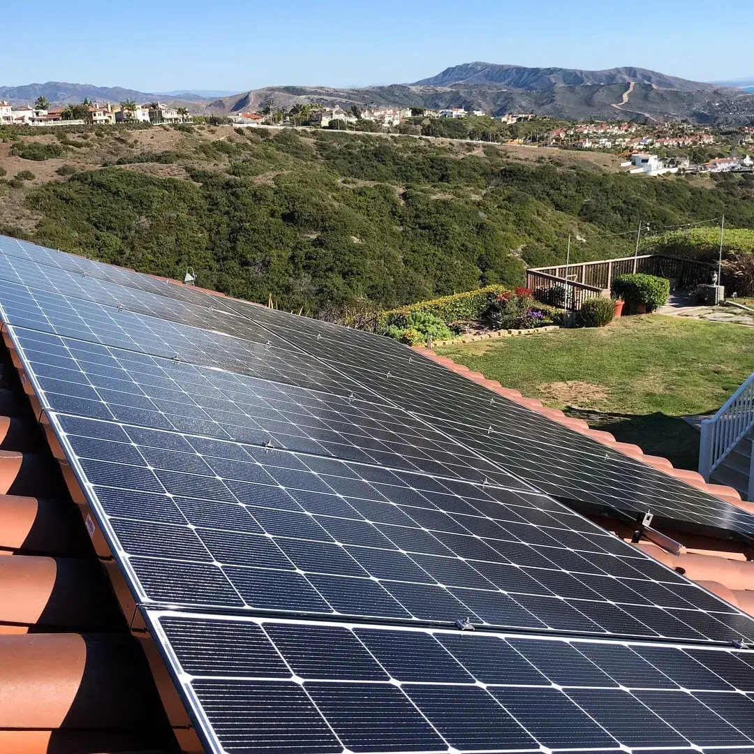 Close-up of solar panels on a tile roof with hills and homes in the background.