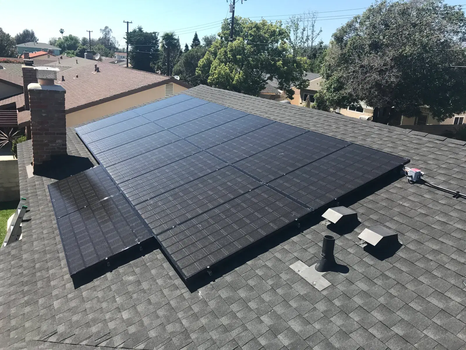 Finished black solar panel array on a gray shingle roof beside a brick chimney under a clear sky.