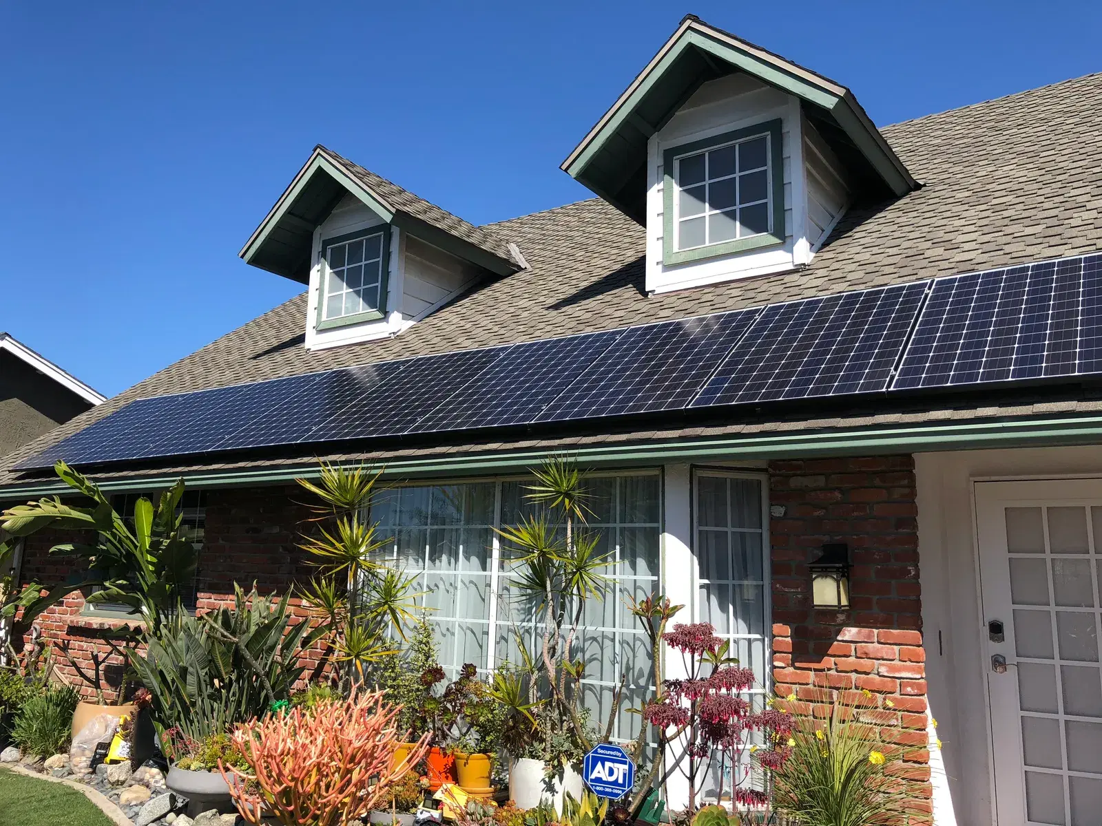 Solar panels on the roof of a single-story brick home with dormer windows and a landscaped garden.
