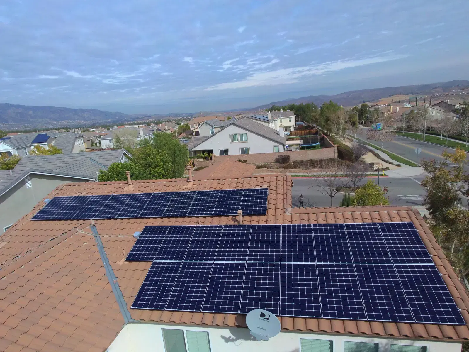 Aerial view of blue solar panels on a red tile roof with a hillside suburban neighborhood beyond.