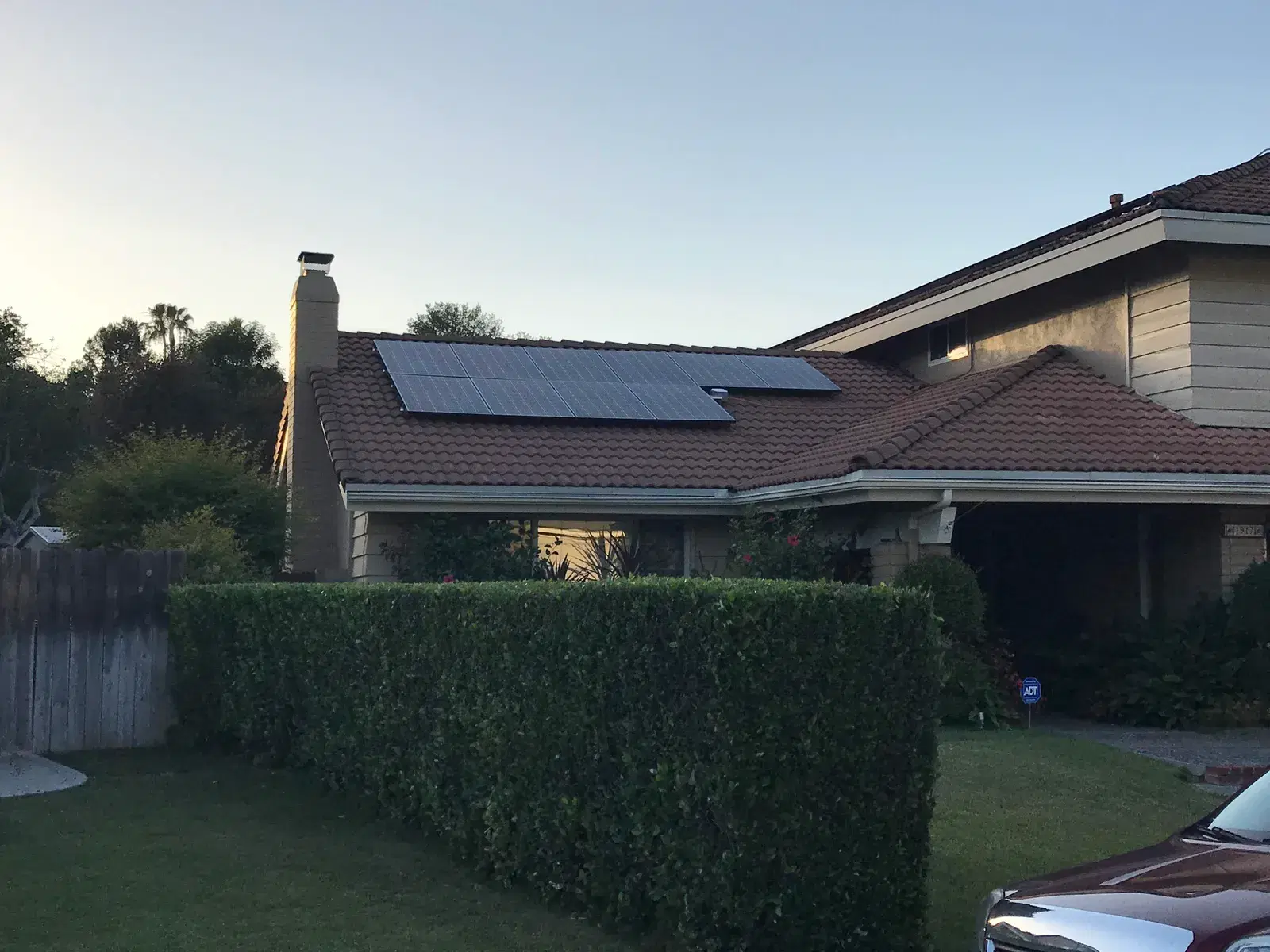 Solar panels on a red tile roof of a two-story home at dusk, viewed over a green hedge from the street.