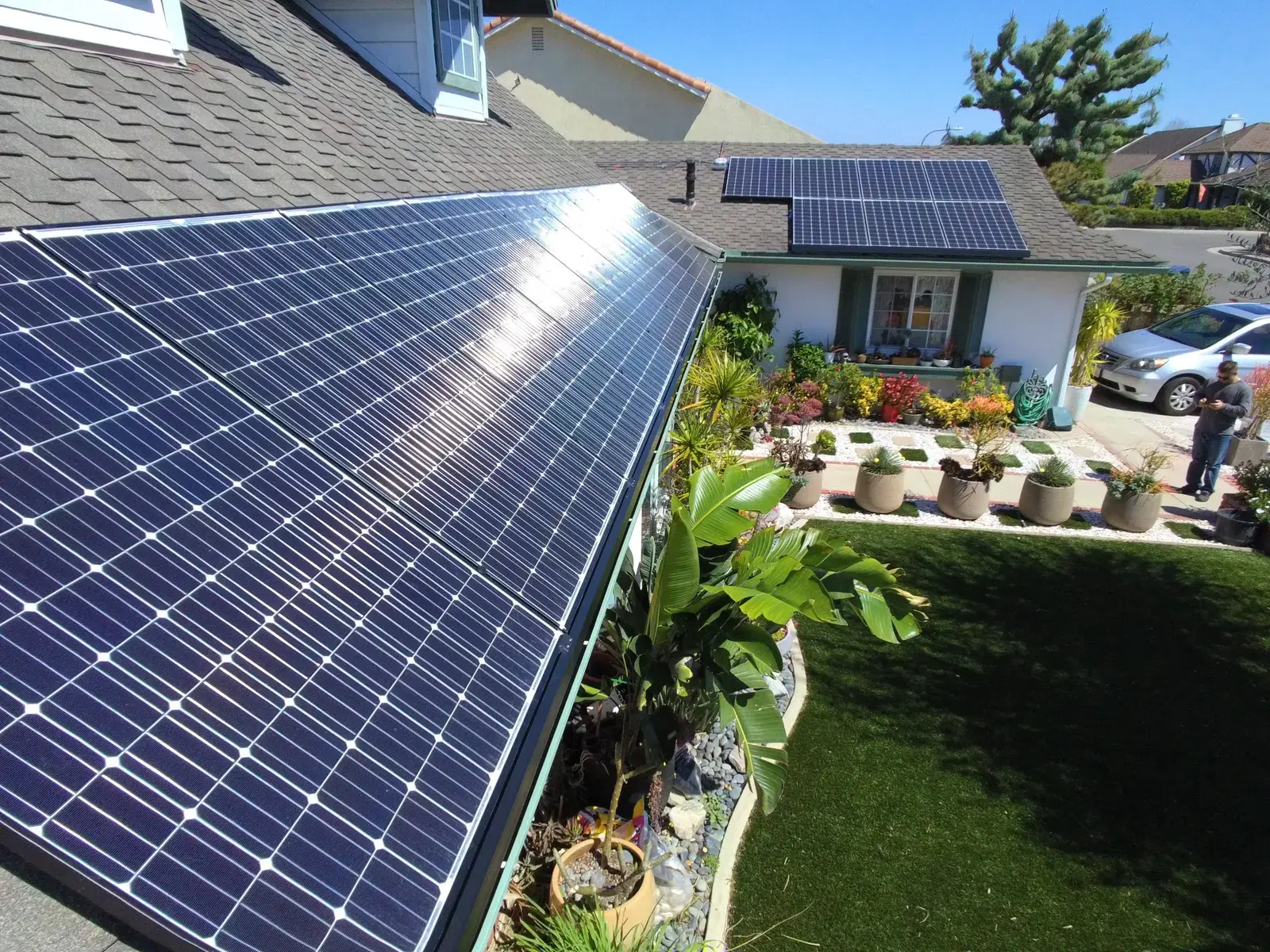 Close-up of monocrystalline solar panels on a sloped residential roof, with a flowering front garden and driveway below.