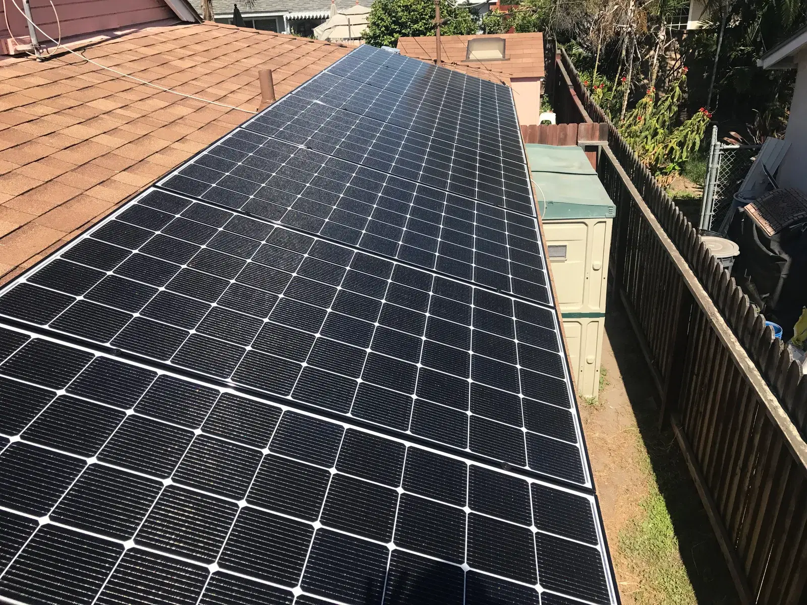 A completed array of black-framed solar panels mounted along a residential roof above a fenced side yard.