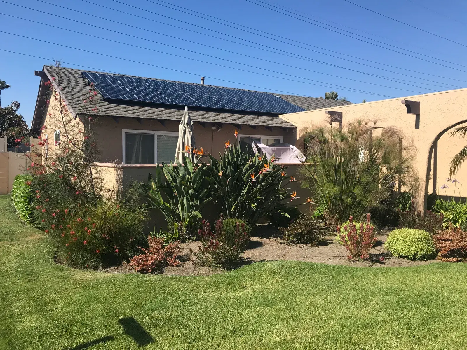 A finished solar array on the roof of a stucco home, viewed across a landscaped backyard with flowers and green lawn.