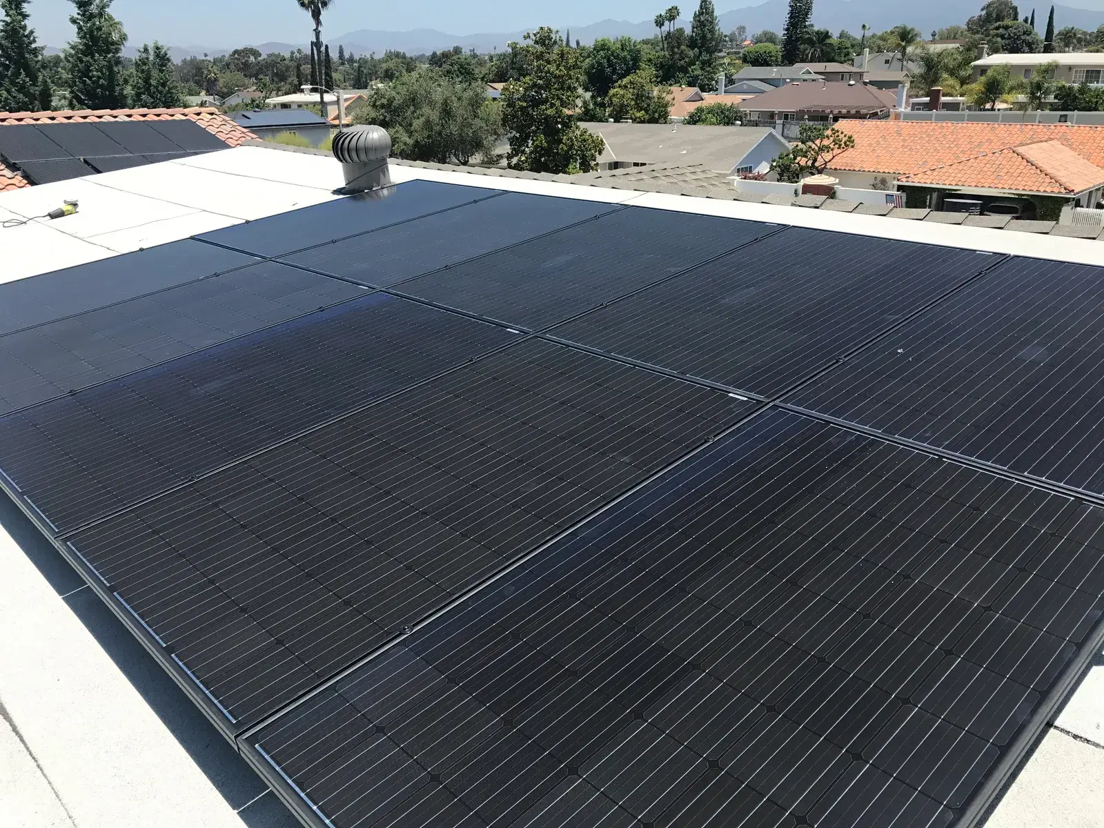 A grid of black rectangular solar panels mounted on a flat white roof, with tile-roofed homes and distant hills in the background.