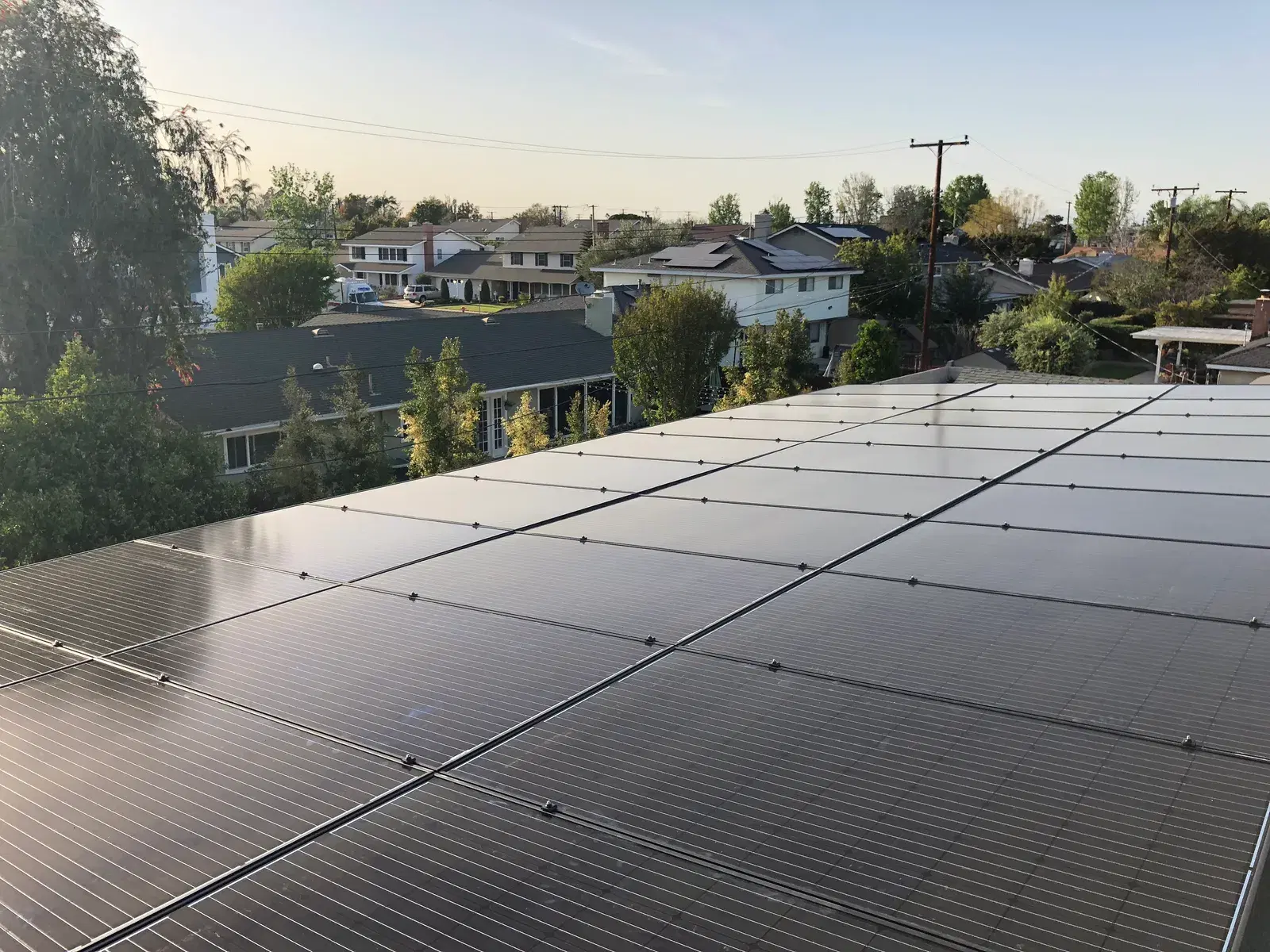 Completed rows of solar panels on a rooftop overlooking single-story homes, trees, and power lines in the soft evening light.