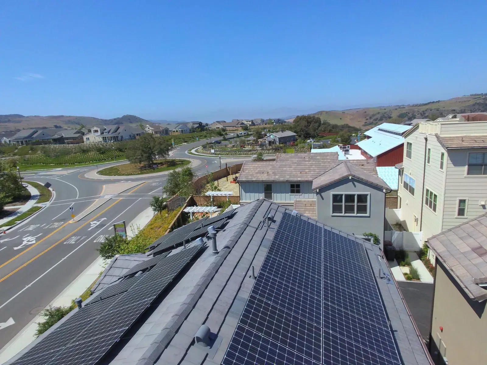 Drone shot of solar panels on a home's roof with a curving suburban street, new homes, and green hillsides under a clear blue sky.