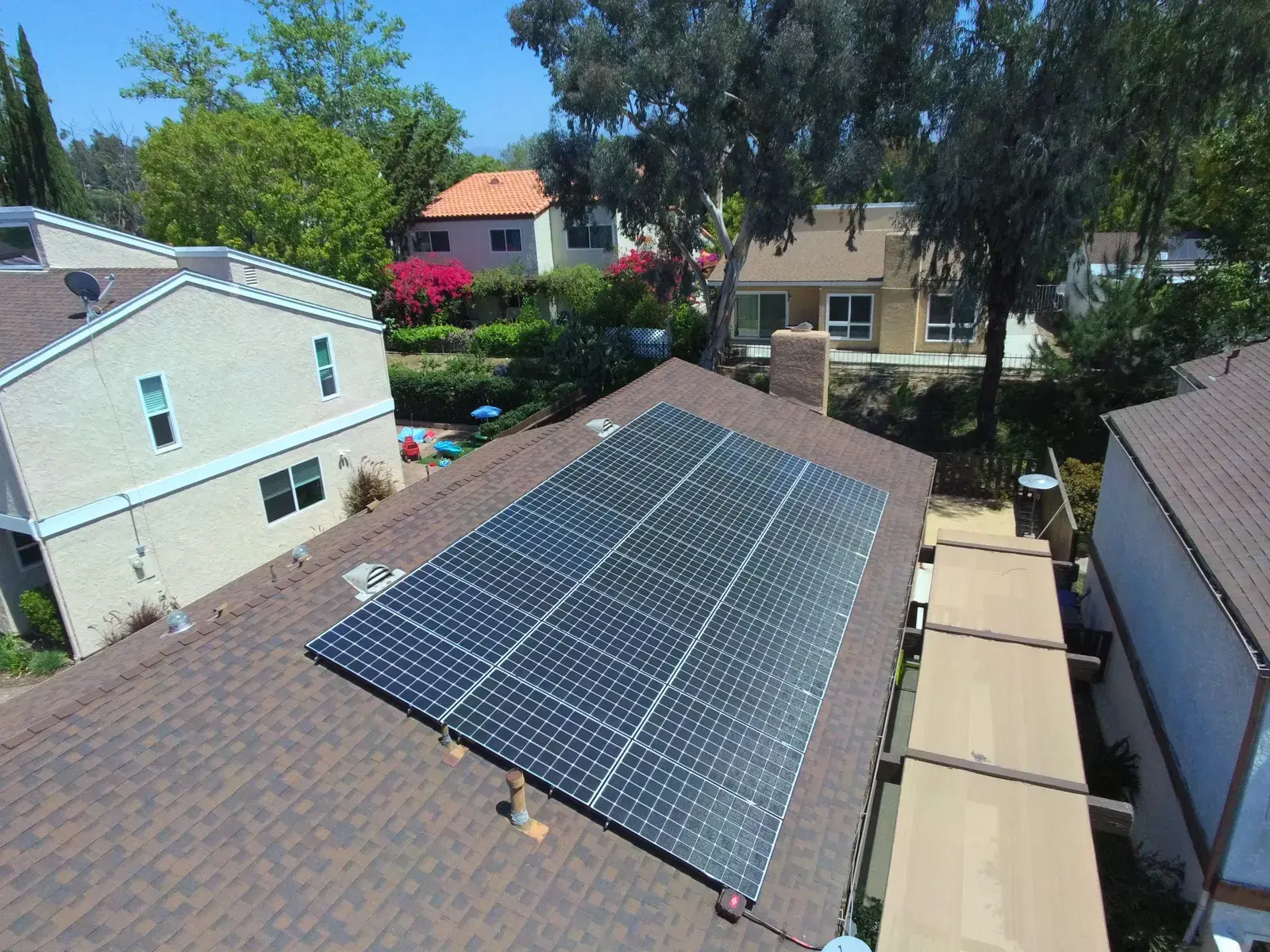 Aerial photo of a residential shingle roof covered with a large grid of solar panels in a green suburban neighborhood.