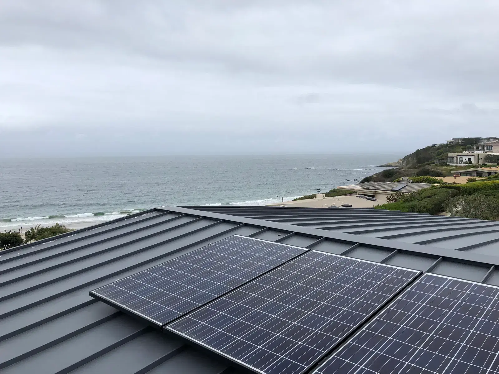 Rectangular photovoltaic panels mounted on a dark metal roof with the ocean and a cliffside beach in the background under overcast skies.