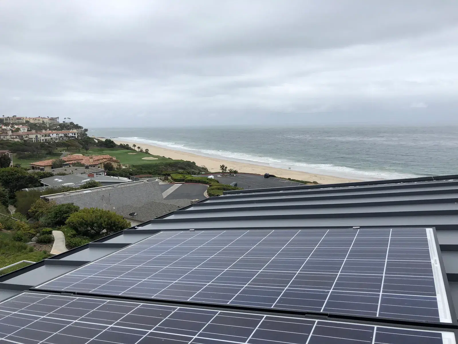 Photovoltaic panels on a dark metal roof with a coastal golf course, sandy beach, and gray ocean stretching to the horizon beyond.