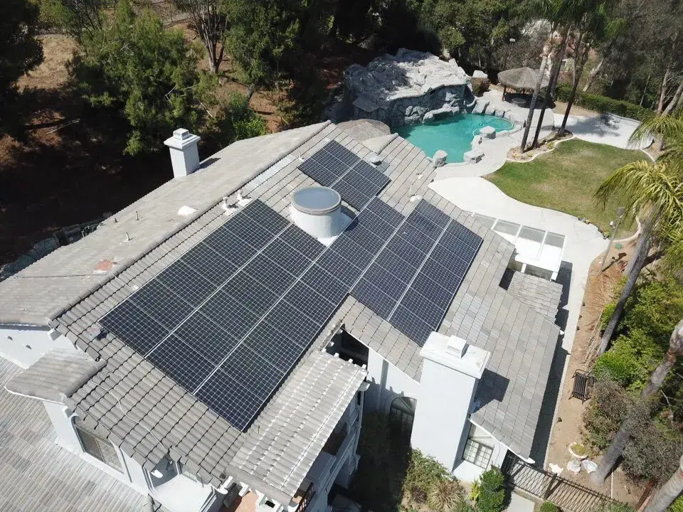 Drone shot looking down on dark solar panels mounted on a gray tile roof, with a backyard pool and lush landscaping beyond.