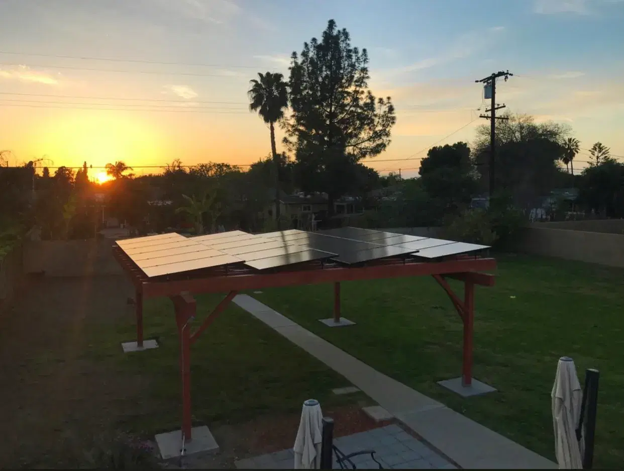 Solar panels mounted on a red-framed freestanding patio cover in a backyard at sunset.