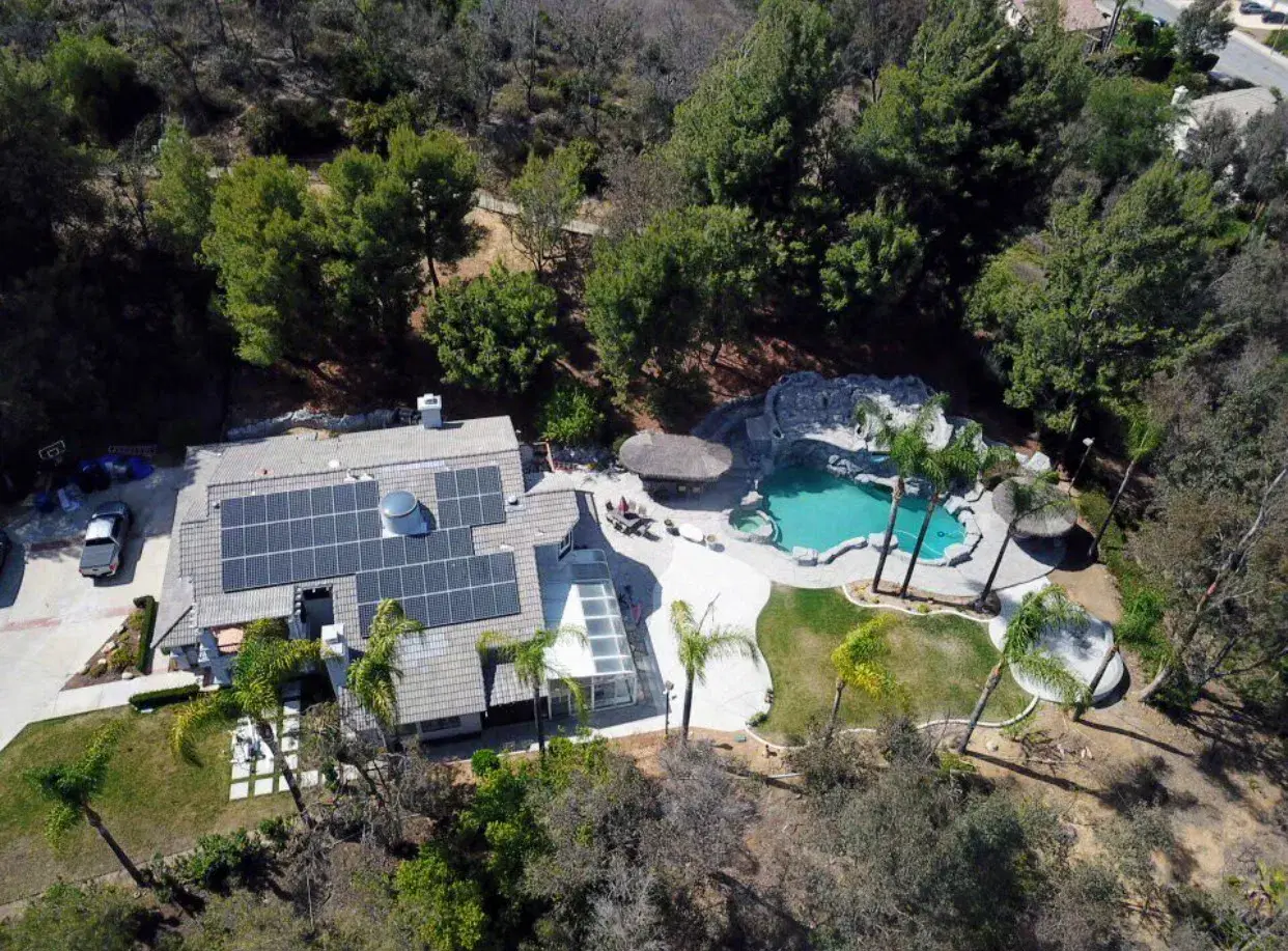 Drone view of a wooded hillside home with rooftop solar panels and a resort-style swimming pool.