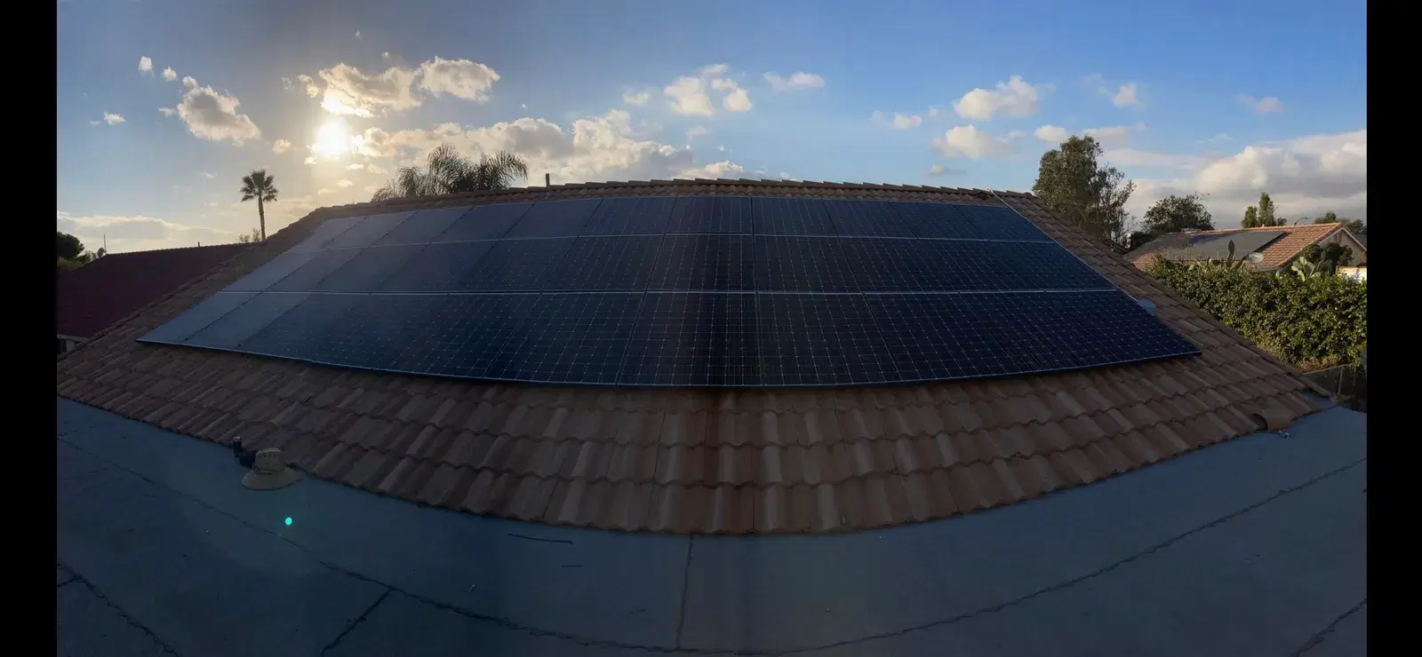 Wide panoramic view of a solar panel array on a tile roof with the setting sun and palm trees in the background.