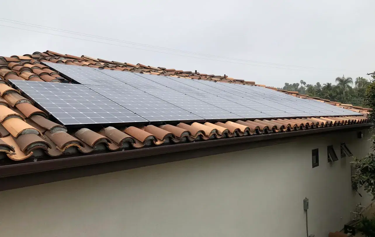 Solar panels installed on a Spanish-style terracotta tile roof of a single-story home, framed by trees on a cloudy day.