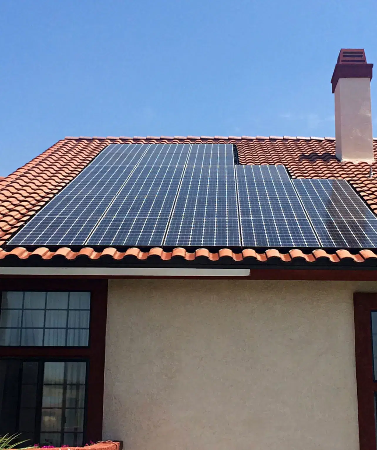 Solar panels mounted on a red clay tile roof beside a stucco chimney under a clear blue sky.