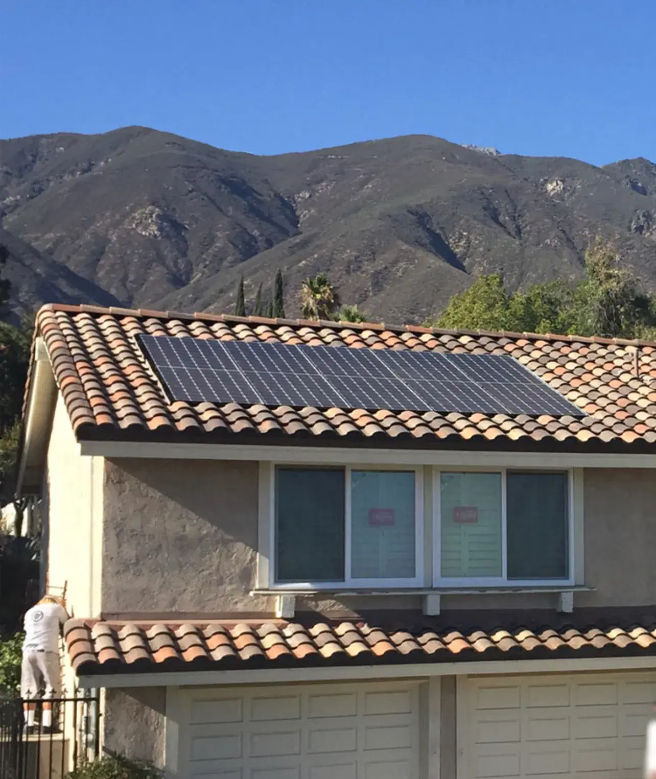 Solar panels on the tile roof of a two-story home with a rugged mountain range rising behind it.