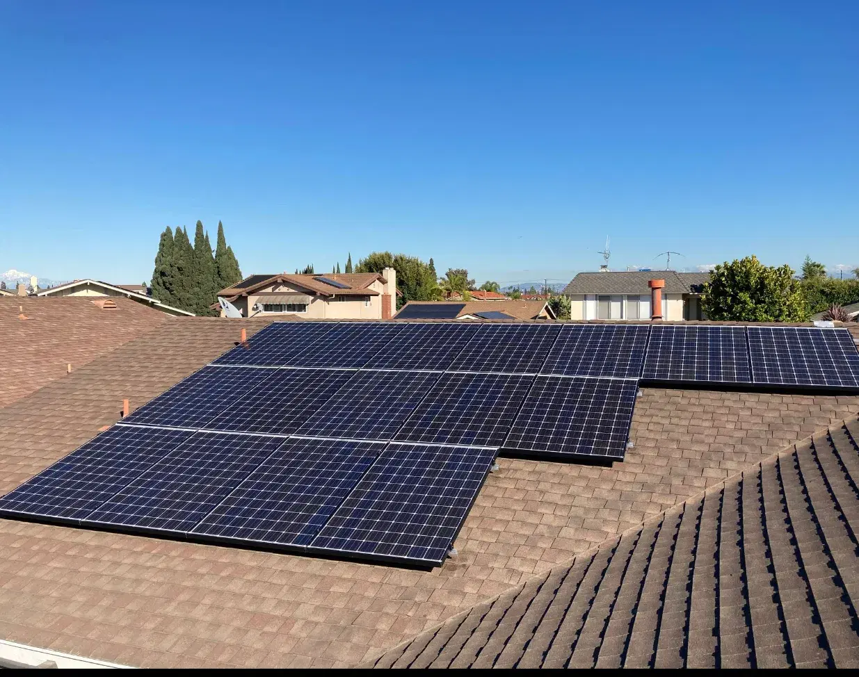 Solar panels installed across a gray shingled roof with neighboring houses and trees in the background.