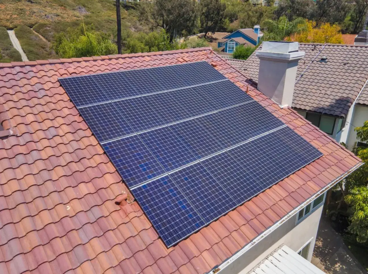 Drone view of solar panels installed on a red clay tile roof with hills and homes in the background.