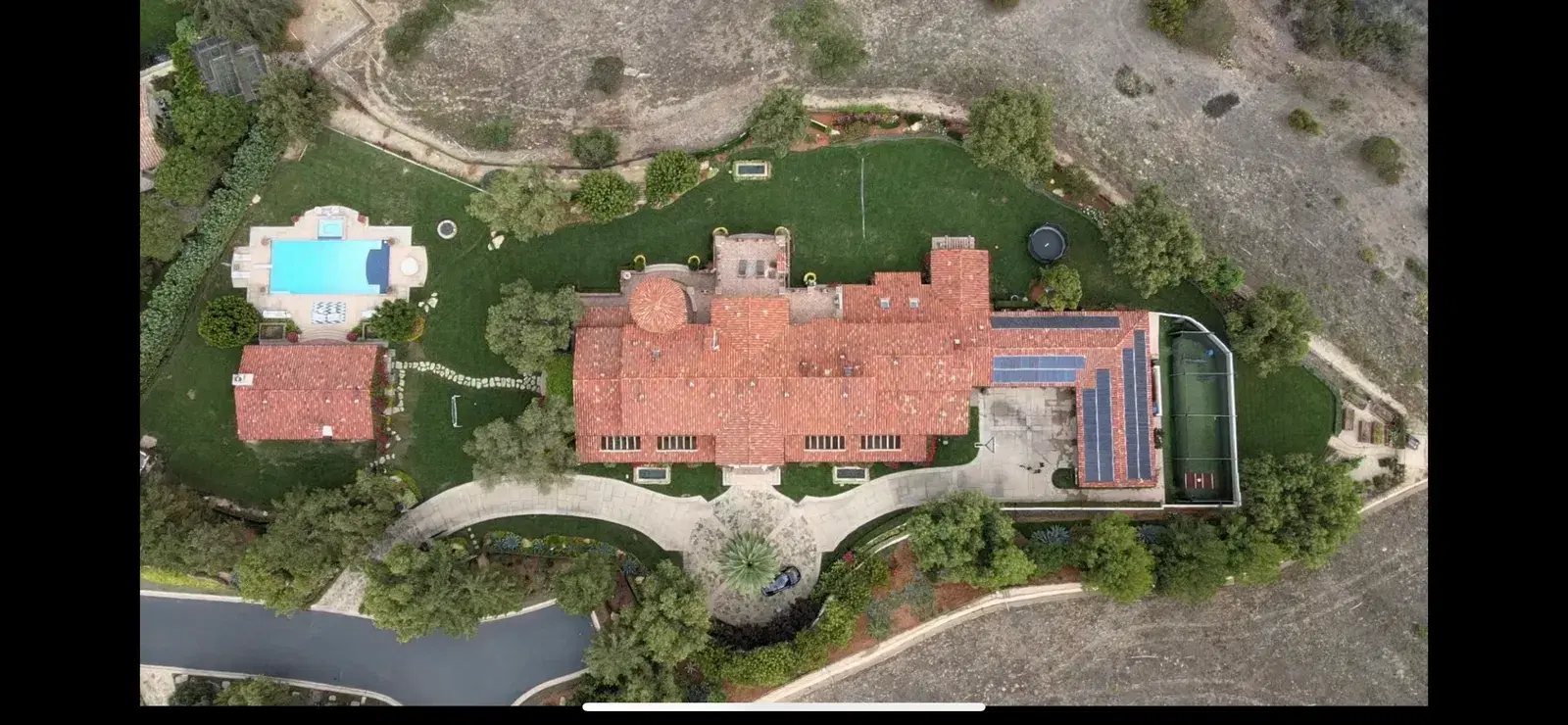 Drone shot looking down on a large red-tile-roofed estate with solar panels, a blue pool, gardens, and a green sport court.