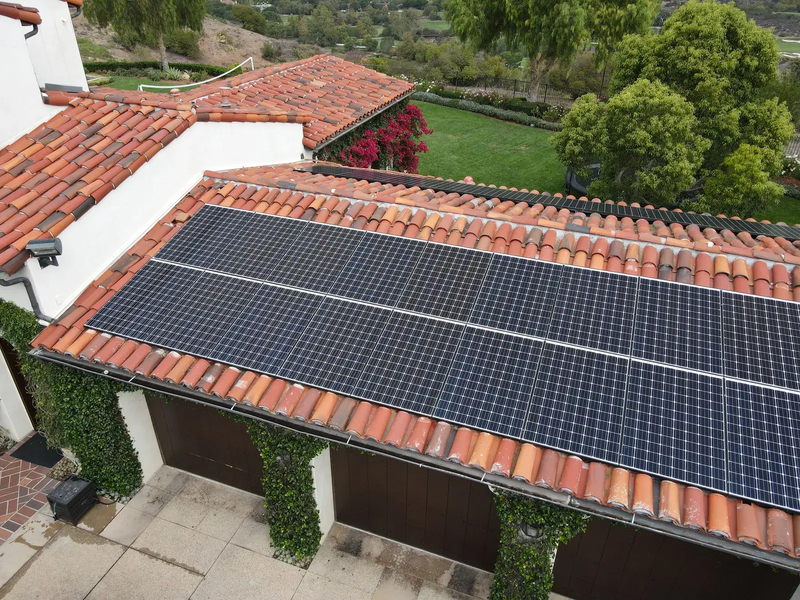 Drone shot of rooftop solar panels installed on a curved terracotta tile roof of a hillside home with a green lawn behind.