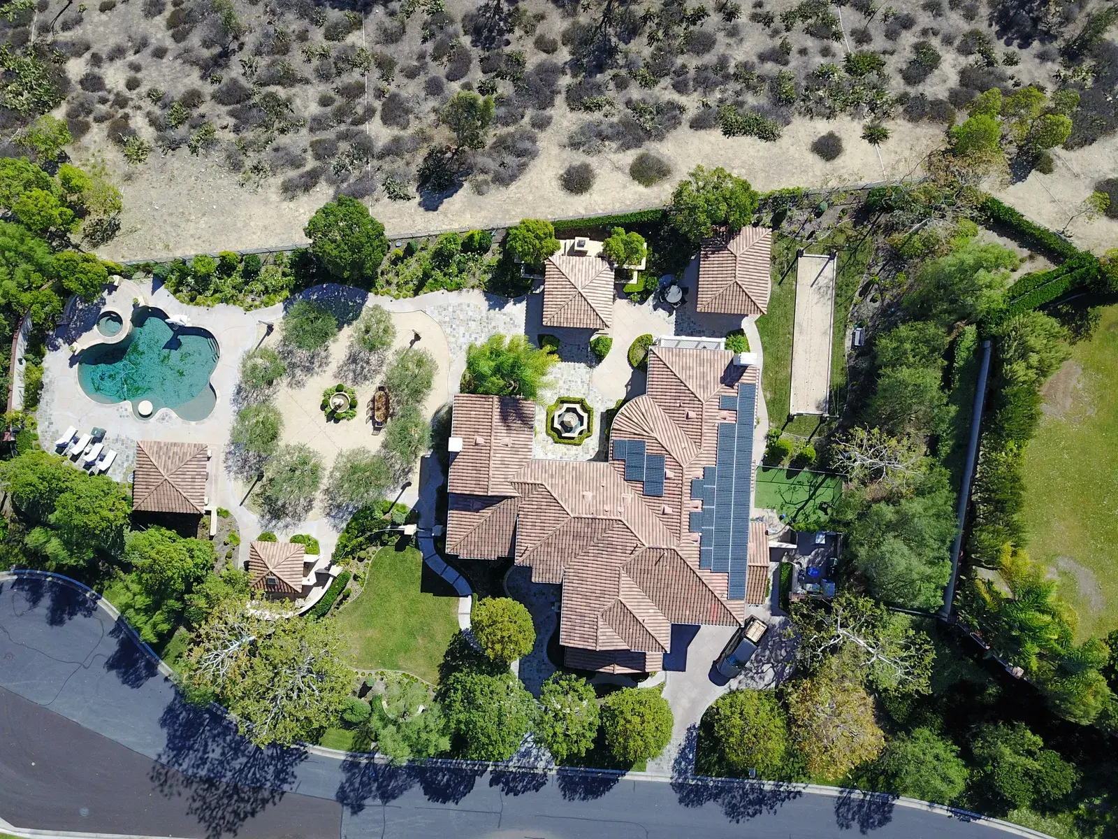 Drone shot looking straight down on a large tile-roof home with black solar panels, a freeform pool and gardens.