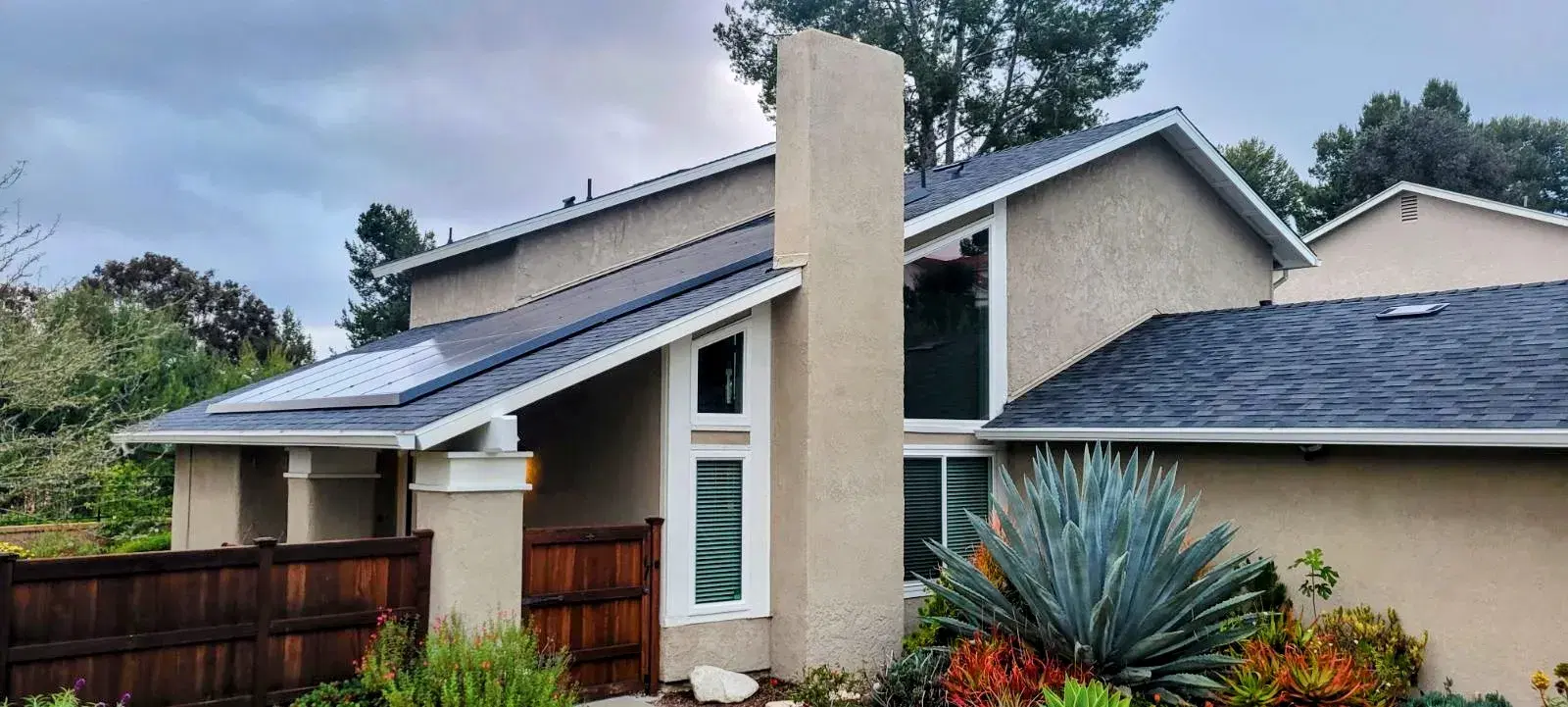 A two-story stucco home with low-profile black solar panels on the sloped roof and agave plants in the foreground.