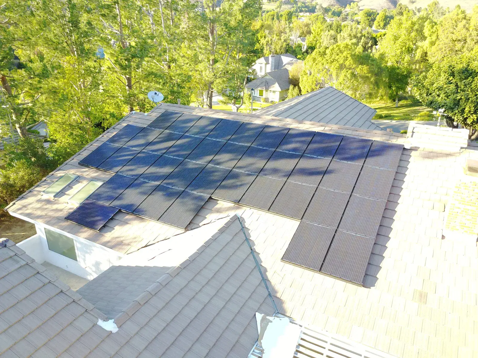Elevated view of black solar panels installed across a tile roof, surrounded by green trees in warm evening light.