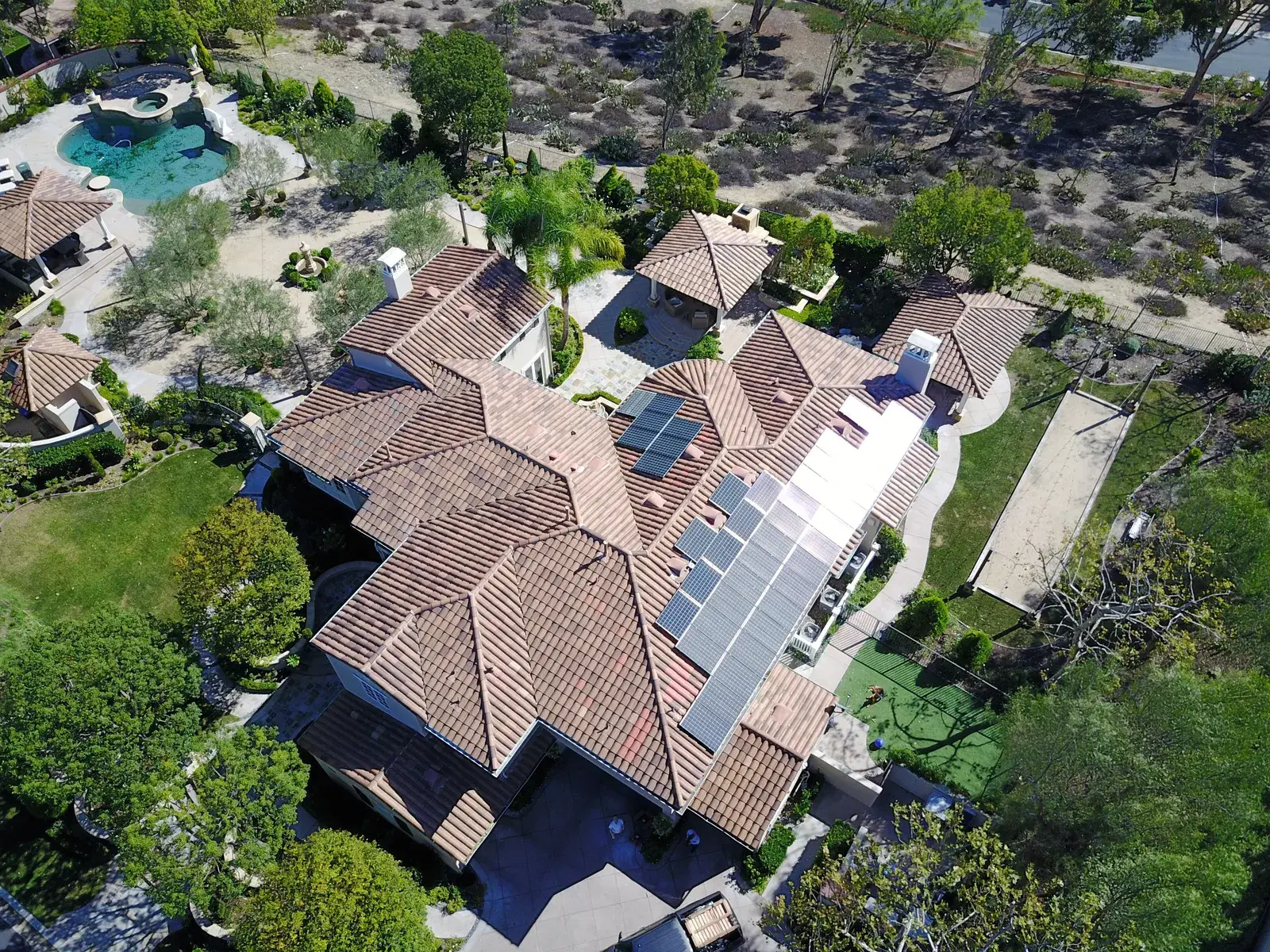 Drone shot of a large terracotta tile-roof home with solar panels, a swimming pool and a tennis court below.